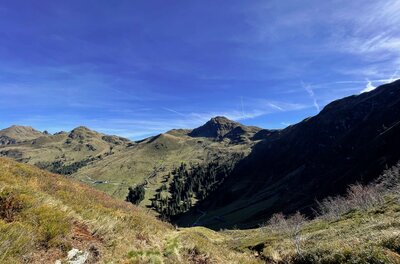 Blick von einem grasbewachsenen Berghang auf gegenüberliegende Berggipfel unter blauem Himmel.