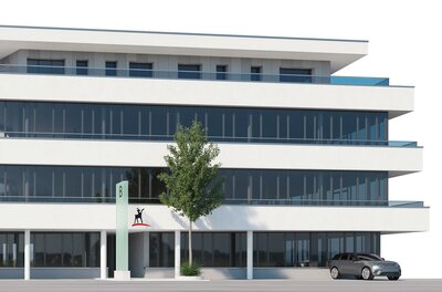 Ein modernes Bürogebäude mit großen Fensterfronten und einem klaren Design. Vor dem Gebäude steht ein Baum und ein Auto parkt auf der Straße.