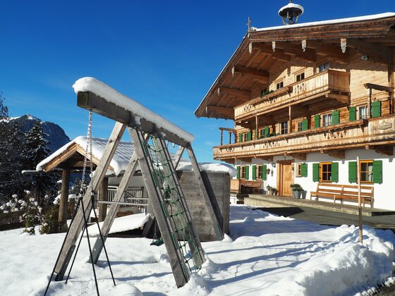 Ein charmantes Chalet im Schnee mit grünen Fensterläden. Neben dem Haus steht eine Schaukel in der verschneiten Landschaft.