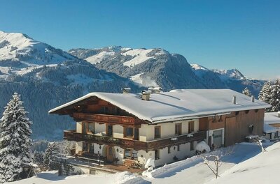 Ein schneebedeckter Bauernhof mit Holzbalkonen vor einer alpinen Bergkulisse im Winter.