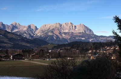 Blick über ein Tal mit Häusern und Wiesen auf ein markantes, felsiges Gebirgsmassiv unter blauem Himmel.
