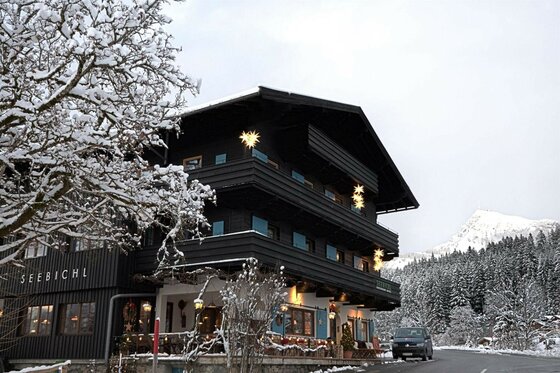 Außenansicht des dunklen Holzgebäudes Seebichl im Winter mit verschneiten Bäumen und Bergen im Hintergrund.