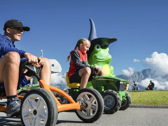 Cart Zwei Kinder fahren mit kleinen Fahrzeugen auf einer sonnigen Wiese. Im Hintergrund ist eine große grüne Figur mit einem Zauberhut zu sehen.