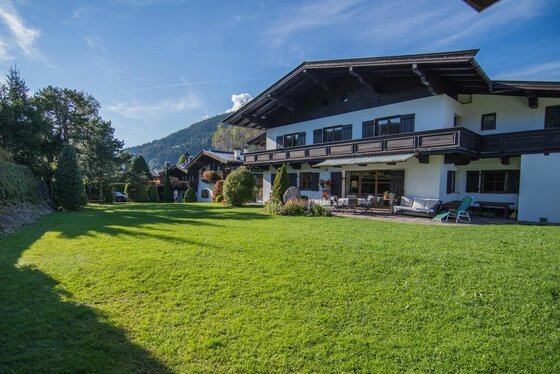 Alpines Landhaus mit großer Rasenfläche, Holzbalkonen und Terrasse unter blauem Himmel.