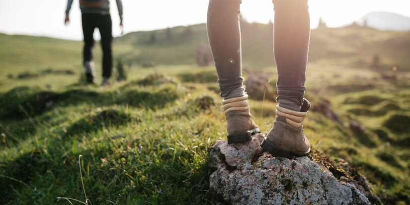 Eine Wanderin mit robusten Stiefeln steht auf einem Stein, während ein anderer Wanderer in der Ferne die Landschaft erkundet.