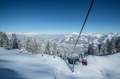 Rote Gondel einer Seilbahn über verschneiten Hängen und Tannen vor Gebirgspanorama.