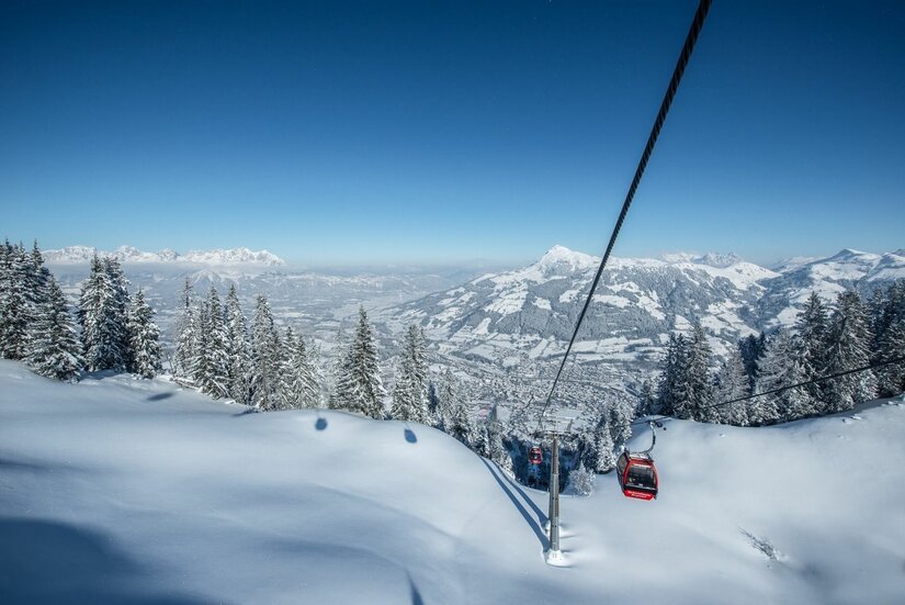 Rote Gondel einer Seilbahn über verschneiten Hängen und Tannen vor Gebirgspanorama.