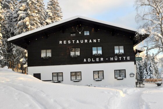 Adlerhütte im Winter Vorderansicht der Adlerhütte im Schnee mit dunkler Holzfassade und weißem Erdgeschoss vor verschneiten Tannen.