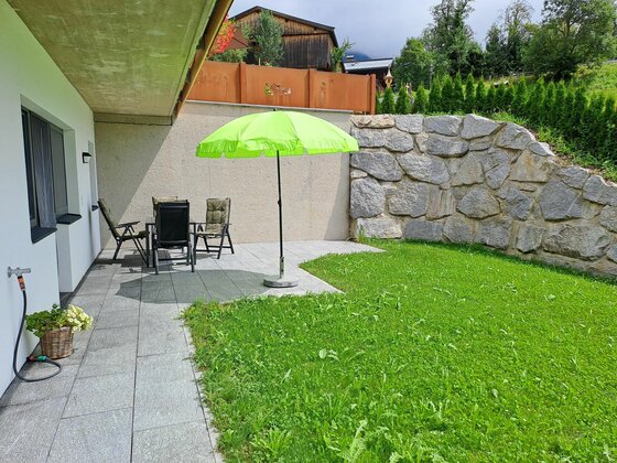 Ferienwohnung Widmann Garten Gepflasterte Terrasse mit Sitzgruppe, grünem Sonnenschirm und angrenzender Rasenfläche vor einer Natursteinmauer.