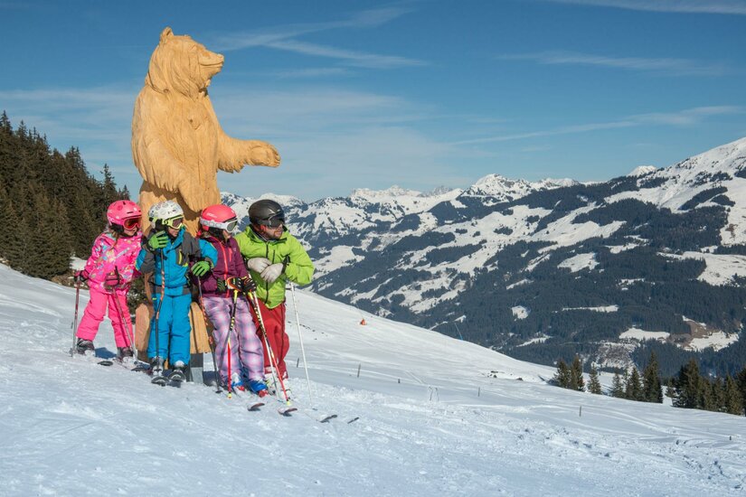 Bärenland Eine Gruppe von Skifahrern in bunten Skianzügen steht im Schnee, umgeben von einer beeindruckenden Berglandschaft. Im Hintergrund steht eine große Bärenfigur.