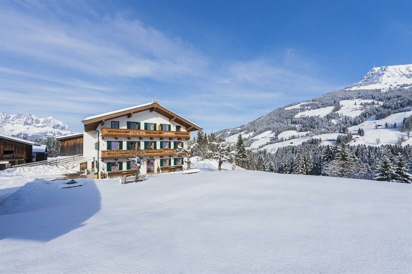 Hof Filzen im Winter Ein traditionelles Alpenhaus mit Holzbalkonen in einer verschneiten Berglandschaft unter blauem Himmel.