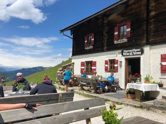 Terrasse der Wiegalm Gäste sitzen an Holztischen vor dem Alpengasthaus Wiegalm unter blauem Himmel.