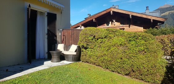 Terrasse mit Rattanmöbeln, kleiner Rasenfläche und Hecke vor einem Holzhaus.
