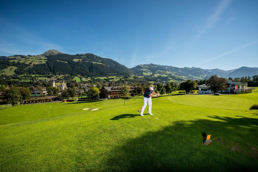 Eine Person beim Golfschlag auf einem grünen Rasenplatz vor einer Bergkulisse.