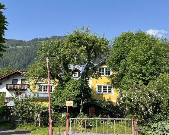 Wohnhaus Schmiedhäusl Gelbes Wohnhaus mit Garten und Metallzaun vor bewaldetem Berghang unter blauem Himmel.