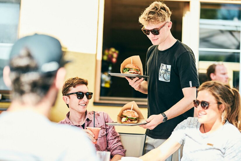 Ein Kellner bringt Burger zu einer Gruppe von Freunden in einem Restaurant. Alle tragen Sonnenbrillen und genießen die Atmosphäre.