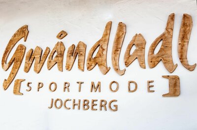 Gwändladl Sportmode Jochberg Holzschriftzug Gwändladl Sportmode Jochberg an einer weißen Wand.
