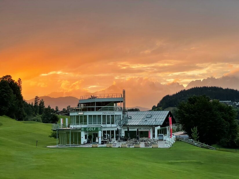 Ein modernes Golfhaus umgeben von grünem Rasen. Im Hintergrund leuchtet der Himmel in warmen Farben bei Sonnenuntergang.