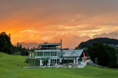 Red Bull Sommer Ein modernes Golfhaus umgeben von grünem Rasen. Im Hintergrund leuchtet der Himmel in warmen Farben bei Sonnenuntergang.