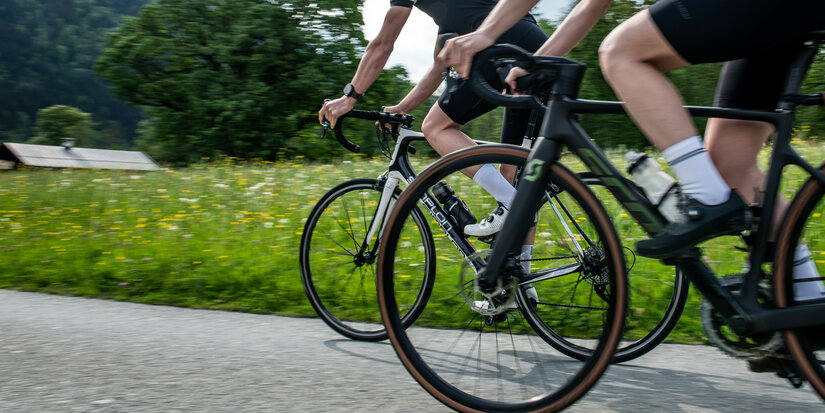 Zwei Radfahrer in sportlicher Kleidung fahren auf einem schmalen Weg, umgeben von blühenden Wiesen und Bäumen.