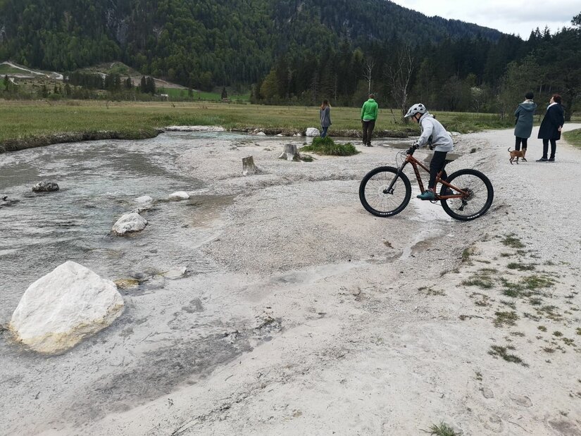 Mountainbiker am Bachlauf Ein Kind auf einem Mountainbike fährt an einem flachen Bachlauf vorbei, im Hintergrund Spaziergänger und Wald.