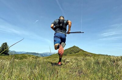 Rückansicht eines Trailrunners mit Laufweste und Stöcken auf einer Bergwiese.