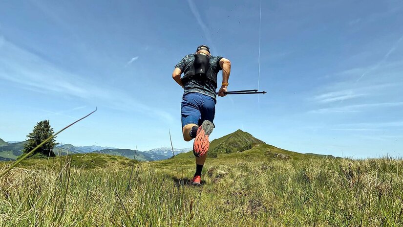 Trailrunning in den Bergen Rückansicht eines Trailrunners mit Laufweste und Stöcken auf einer Bergwiese.