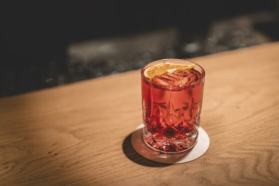 Ein Glas mit rotem Getränk, Eiswürfeln und einer getrockneten Orangenscheibe auf einem Holztisch.