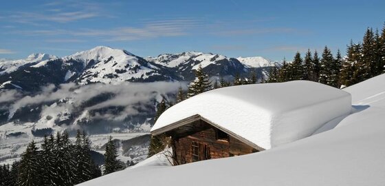 Holzhütte mit dicker Schneeschicht auf dem Dach vor verschneitem Bergpanorama und Nadelwald.