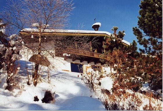 Alpines Landhaus mit Holzbalustrade im verschneiten Hang unter blauem Himmel.