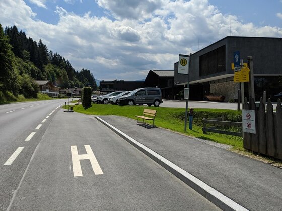 Bushaltestelle an einer Landstraße mit Wartebank, Haltestellenschild und Parkplatz vor modernen Gebäuden.