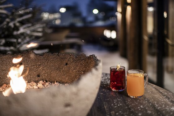 LaMonte Bistro Außenbereich Zwei Gläser mit Heißgetränken neben einer brennenden Feuerschale auf einer winterlichen Terrasse am Abend.
