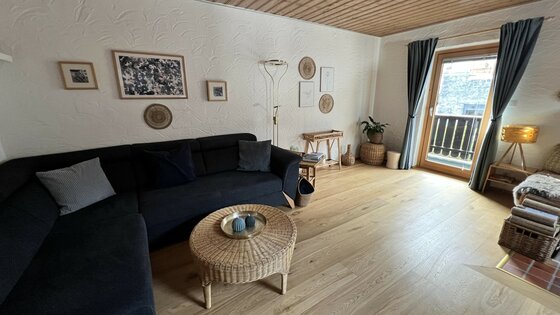 Ein gemütliches Wohnzimmer mit einem dunklen Sofa und einem runden Tisch. Durch die Fenster scheint Tageslicht und es gibt einen Zugang zu einem Balkon.