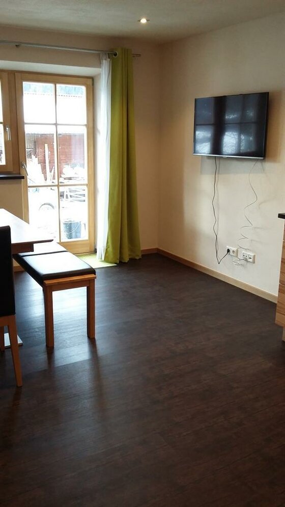 Wohnbereich mit dunklem Boden, Esstisch, Sitzbank und wandmontiertem Fernseher.