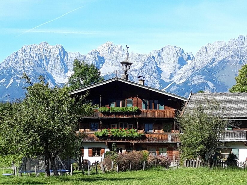Traditionelles Holzhaus mit Blumenschmuck vor einer massiven, schneebedeckten Gebirgskette unter blauem Himmel.