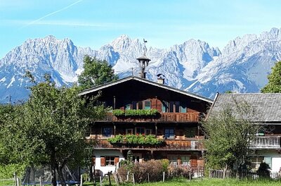 Traditionelles Holzhaus mit Blumenschmuck vor einer massiven, schneebedeckten Gebirgskette unter blauem Himmel.