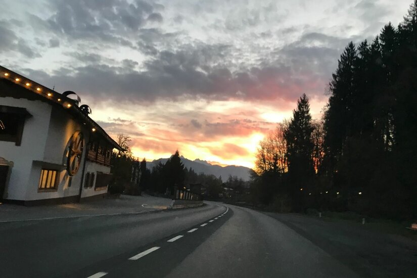 Asphaltierte Landstraße bei Sonnenuntergang mit einem alpinen Gebäude am linken Rand und Wald rechts.