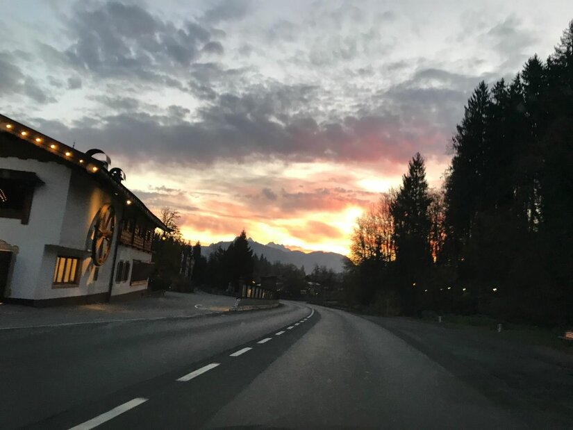 Landstraße bei Sonnenuntergang Asphaltierte Landstraße bei Sonnenuntergang mit einem alpinen Gebäude am linken Rand und Wald rechts.