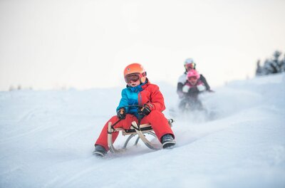 Rodeln am Gaisberg Ein Kind mit Helm und Skibrille fährt auf einem Holzschlitten eine verschneite Piste hinunter.