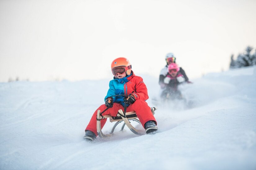 Ein Kind mit Helm und Skibrille fährt auf einem Holzschlitten eine verschneite Piste hinunter.