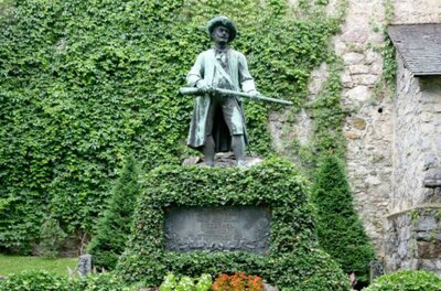 Freiheitskämpferdenkmal Eine Bronzestatue eines Mannes mit Gewehr steht in einem grünen Garten. Der Hintergrund ist von Efeu und einer Steinmauer umgeben.