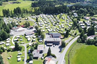 Luftaufnahme eines Campingplatzes mit zahlreichen Wohnwagen, Gebäuden und angrenzendem Wald am See.