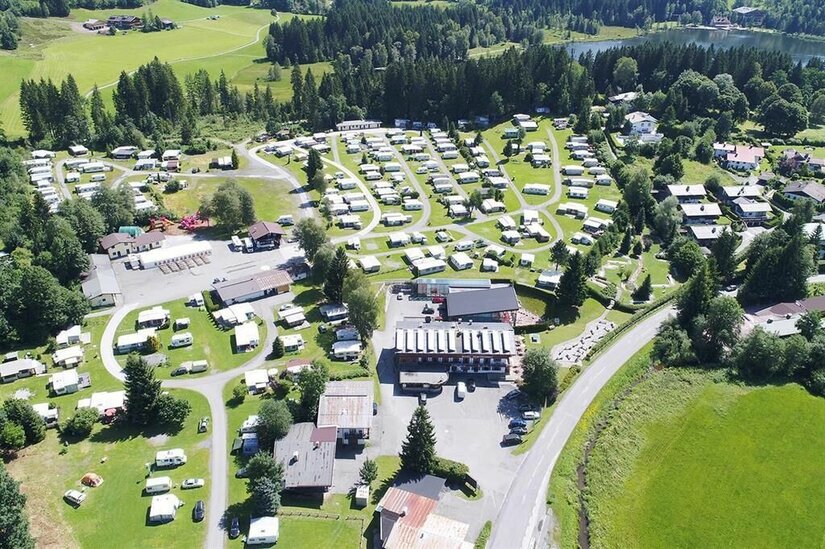 Luftaufnahme eines Campingplatzes mit zahlreichen Wohnwagen, Gebäuden und angrenzendem Wald am See.