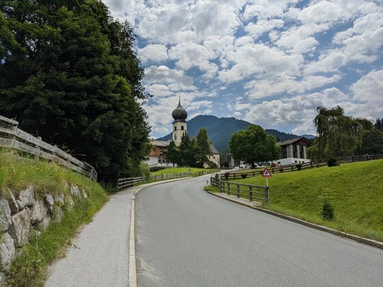 Asphaltierte Straße führt auf eine Kirche mit Zwiebelturm und bewaldete Berge unter bewölktem Himmel zu.