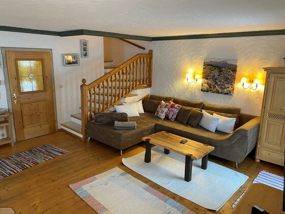 Wohnbereich Haus Staudach Wohnzimmer mit braunem Ecksofa, Holztisch und einer Holztreppe im Hintergrund.