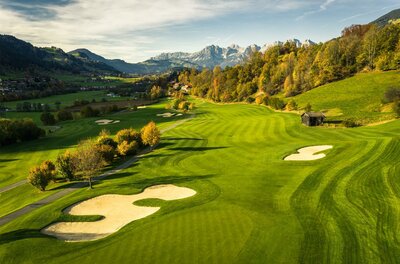 Eine malerische Golfanlage mit grünen Fairways und Bunkern. Im Hintergrund erheben sich majestätische Berge und ein klarer Himmel.