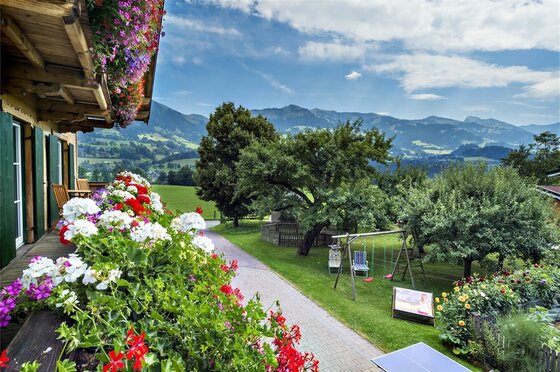 Blick von einem blumengeschmückten Holzbalkon auf einen Garten mit Spielplatz und Berge im Hintergrund.