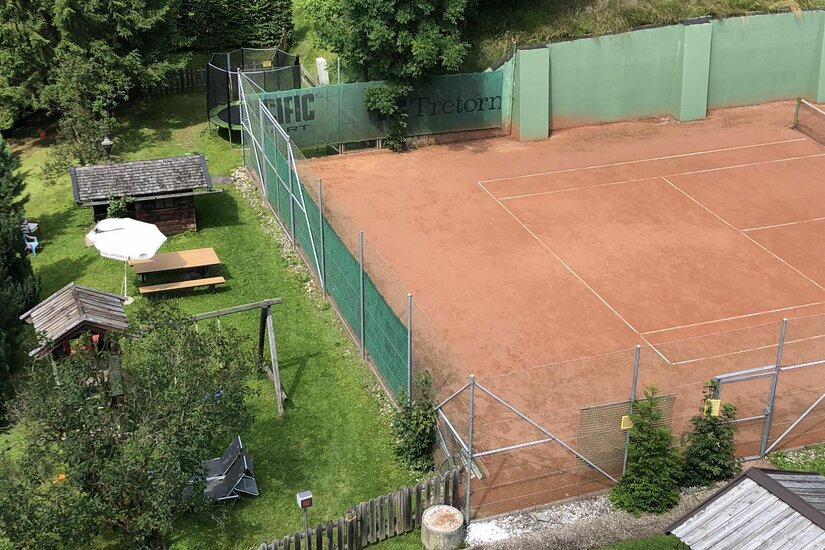 Ein Tennis-Sandplatz neben einer Liegewiese mit Spielplatz und Trampolin.