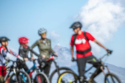 Vier Mountainbiker stehen mit Helmen vor einer Bergkulisse mit Sendeturm.