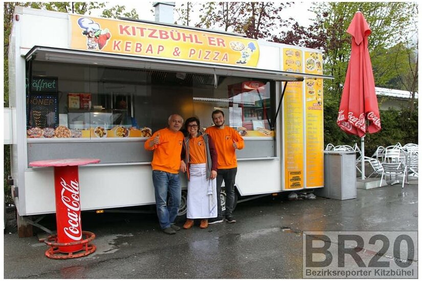 Ein Kiosk in Kitzbühel, der Kebap und Pizza verkauft. Drei Personen in orangefarbener Kleidung posieren freundlich vor dem Verkaufsstand.